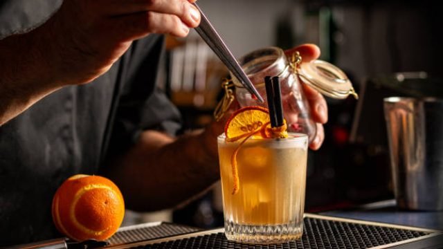 Preparing Smoky Spiced Orange Cocktail decorated with Dehydrated Orange Slices anf fresh orange peel. Bar or pub interior.